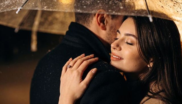 Romantic couple embracing under clear umbrella at night showing engagement ring