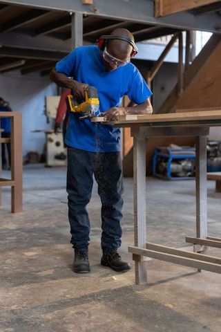 African american carpenter using hand router cutting wood plank in industrial workshop
