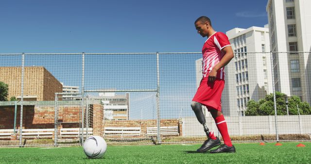 A young soccer player with a prosthetic leg practices controlling a ball on an outdoor soccer field surrounded by tall buildings on a bright, clear day. This image can be used to depict themes of determination, resilience, disabled sports, inclusivity in athletics, and overcoming challenges.