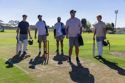 Diverse cricket teammates standing on crease holding bats by stumps on sunny field