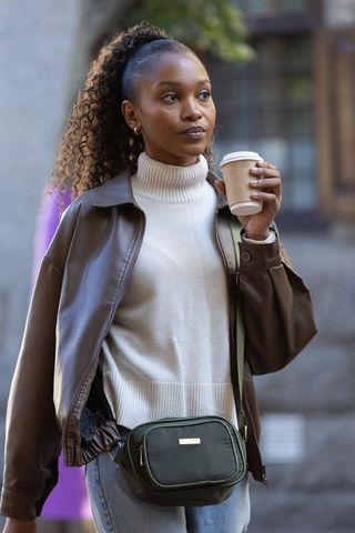 Woman walking urban street holding coffee cup wearing leather jacket and green crossbody bag
