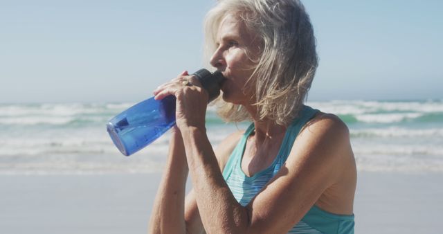 Elderly woman enjoying a sunny day at the beach while staying hydrated by drinking water from a bottle. This image can be used for promoting senior health, outdoor activities, summer vacations, healthy lifestyles, and fitness routines.