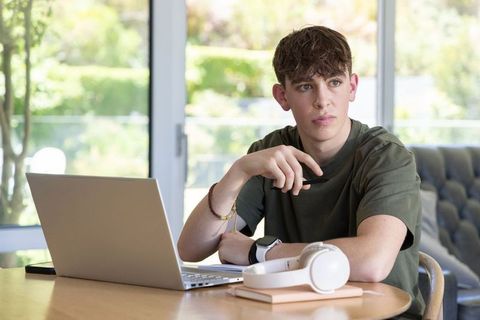 Teenage male studying on laptop at home holding pen with headphones on table