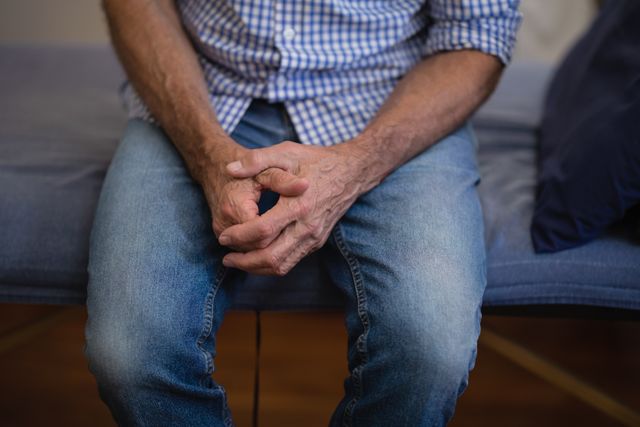 Midsection of senior male patient sitting with hands clasped on bed at hospital ward