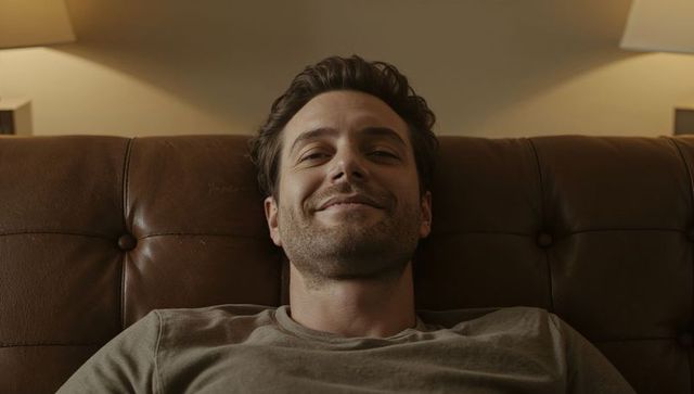 Relaxing young man reclining on brown leather sofa under warm lamp light, cozy evening