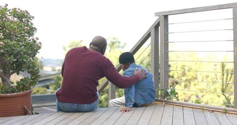 African american father comforting son on deck overlooking hillside with greenery