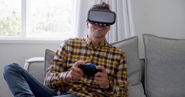 This image shows a man smiling while using a VR headset and holding a game controller, sitting casually on a couch in a living room. Perfect for illustrating home entertainment, modern technology, gaming, and relaxation. Ideal for use in articles, blogs, advertisements related to VR gaming, tech products, and leisure activities.