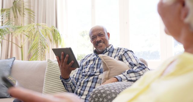 Elderly man sitting on a couch using a digital tablet, appearing relaxed and content. The scene is set in a bright and cozy living room with natural light and plants in the background. This image can be used for themes related to senior lifestyle, technology adaptation, healthy aging, leisure activities, and family moments.