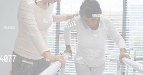 Caregiver helping mature woman practicing gait on parallel bars in rehabilitation clinic