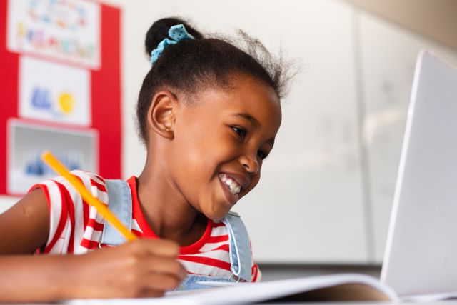 Smiling african american elementary schoolgirl using laptop while writing on book at desk in class. unaltered, education, learning, happiness, wireless technology, studying and school concept.