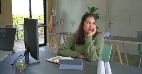 Designer daydreaming at modern home office desk with blueprints, tablet and green sweater