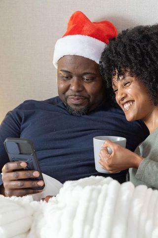 Couple cuddling on couch wearing santa hat sharing coffee under chunky knit blanket