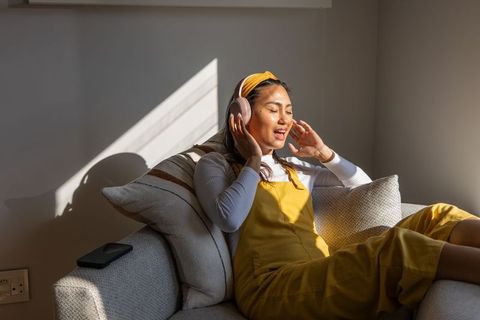 Asian woman relaxing on sofa listening to music with pink headphones in sunlit room
