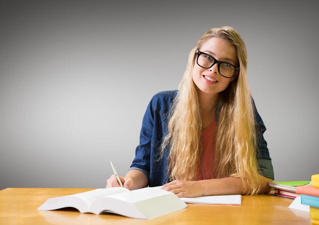 Young woman with glasses sitting at work desk, taking notes with a pencil in hand. Open book in front, with more books on side. Grey background suggesting corporate or educational setting. Could be used for educational content, corporate training materials, and website banners emphasizing study or focus.