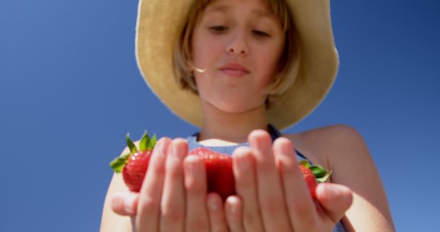 This photo depicts a child holding several fresh strawberries in their hands on a sunny day. The child is wearing a hat and appears to be enjoying the outdoors. This can be used for promoting healthy eating, organic produce, summer activities, or nature-related content.