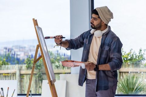 Bearded artist painting on easel in sunlit studio wearing headphones holding paint tray