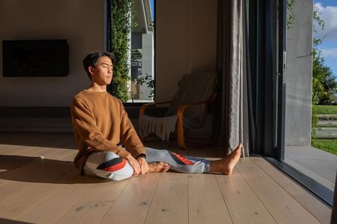 Asian man meditating and stretching in morning sunlight by open sliding glass door