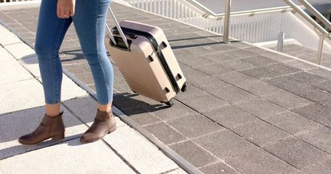 Woman walking pulling beige suitcase up paved ramp wearing skinny jeans and brown boots