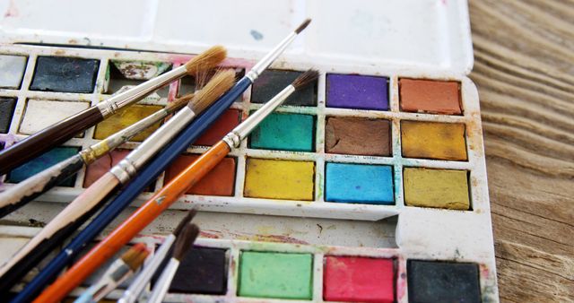 A well-used watercolor palette with various brushes suggests an artist's workspace, with copy space. Vibrant colors and the presence of paintbrushes evoke the creative process and artistic expression.