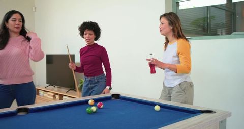 Diverse women playing pool and socializing around blue-felt table in modern home lounge