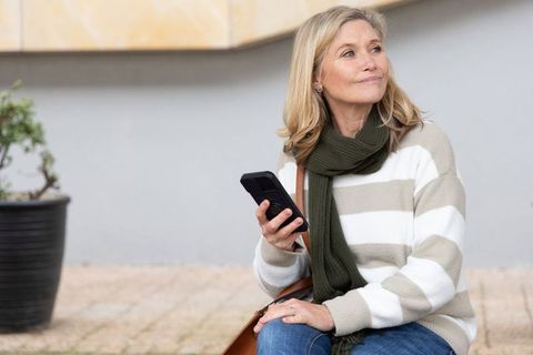 Middle-aged woman checking smartphone while sitting on city bench in casual outfit
