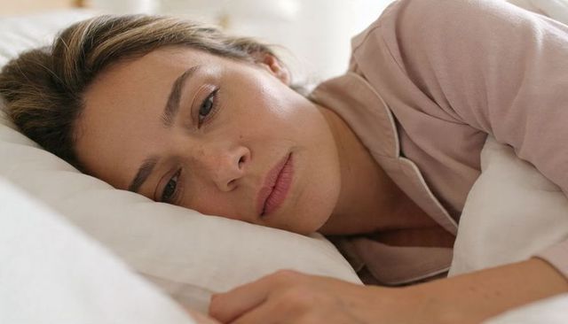 Pensive woman lying in bed wearing light pink pajamas, tired morning close-up portrait