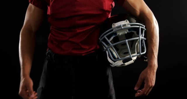 This image depicts a football player holding a helmet against a dark background, highlighting the intensity and readiness of the athlete. Ideal for use in sports-related articles, athletic gear promotions, fitness motivations, team spirit advertisements, and materials aimed at football fans.