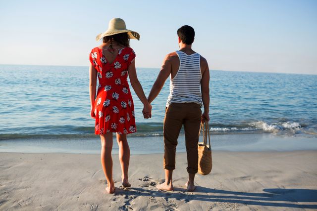 Young couple standing on beach holding hands, enjoying sunny day by the ocean. Ideal for travel brochures, romantic getaway promotions, summer vacation advertisements, and lifestyle blogs focusing on relationships and leisure activities.