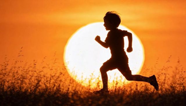 Child running silhouette against oversized sun at sunset over meadow during golden hour