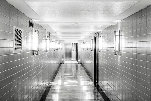 This image features a long, empty corridor with symmetrical fluorescent lighting and tiled walls in a black and white design. Ideal for illustrating concepts of emptiness, solitude, modern architecture, and minimalist design. Useful for websites, architectural blogs, office or school settings, and articles on interior design or institutional environments.