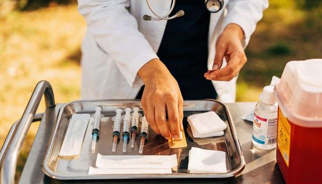 Clinician preparing prefilled syringes on sunlit outdoor vaccination cart with sharps bin