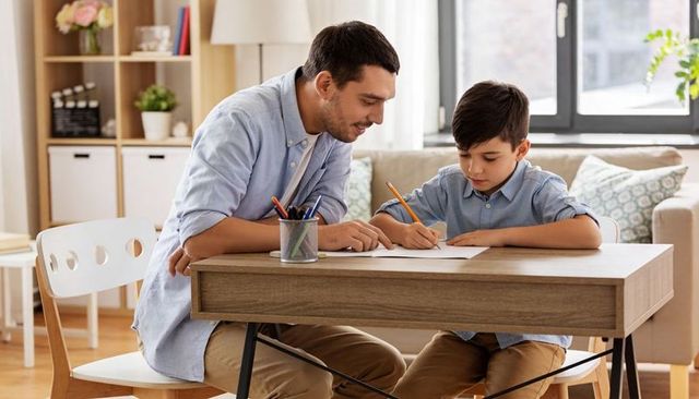 Father tutoring son at home desk, guiding homework and learning in bright living room