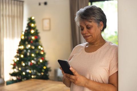 Senior woman using smartphone near christmas tree at home