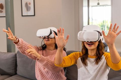 Diverse women laughing and reaching while wearing white vr headsets on gray sofa