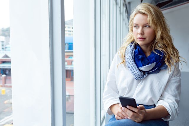 Front view of beautiful young Caucasian businesswoman looking away while using mobile phone near window in a modern office