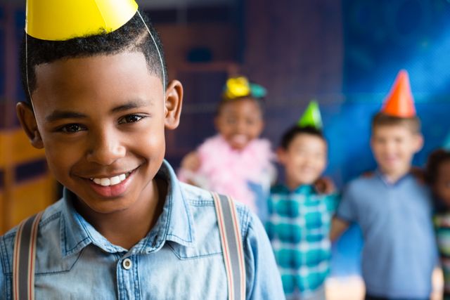 Boy smiling and wearing a party hat with friends in the background during a birthday party. Ideal for use in advertisements, birthday invitations, children's party planning materials, and social media posts celebrating children's events.
