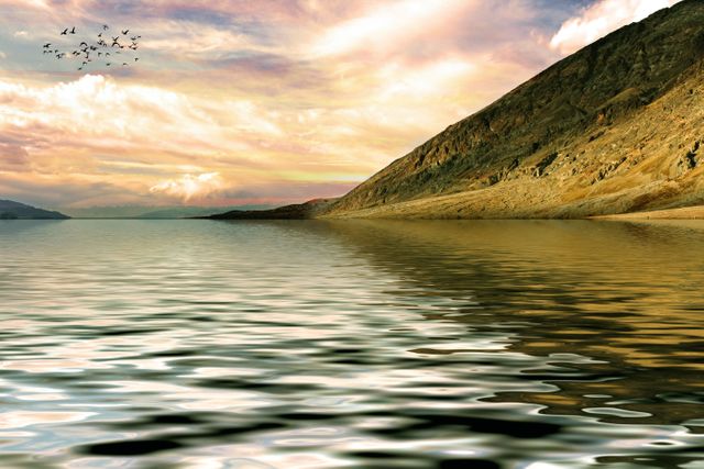 Captivating scene of a tranquil lake at sunset, framed by rugged mountains and calm, reflective water. Birds are seen flying across the sky, adding to the serene atmosphere. Great for use in nature-themed presentations, relaxation backgrounds, and travel advertisements, highlighting tranquility and natural beauty.