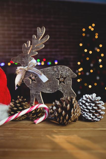 Rustic Christmas decorations featuring a wooden reindeer, pinecones, candy cane, and Santa hat on a wooden table. Blurred Christmas lights and tree in the background add a festive touch. Ideal for holiday greeting cards, seasonal promotions, and festive home decor inspiration.