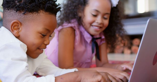 Two African American children lying on the floor and using a laptop together. Both kids are smiling and engaged, enjoying their time together with technology. Could be used for themes like family bonding, digital learning, education from home, online learning, technology in education, or childhood happiness.