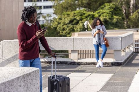 Young black traveler checking phone while wheeling suitcase on urban rooftop terrace