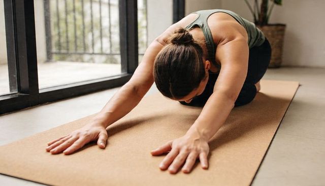 Woman practicing child's pose on cork yoga mat near glass doors in sunlit morning studio