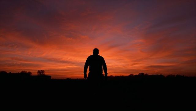 Male silhouette standing against fiery sunset over rural field at twilight, dramatic sky