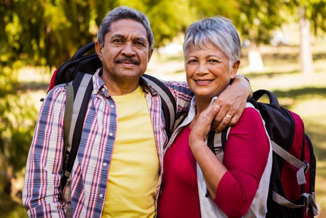 Senior couple embracing while hiking in a park, both wearing backpacks and smiling. Ideal for use in advertisements promoting active lifestyles, retirement activities, outdoor adventures, and healthy living for mature adults. Perfect for travel brochures, health and wellness campaigns, and senior community promotions.