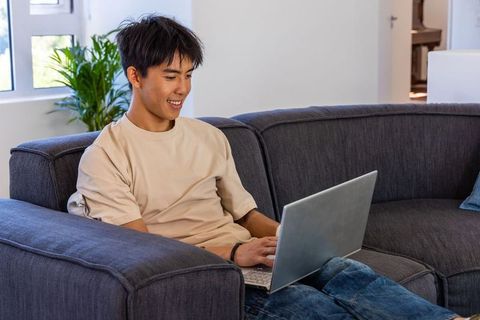 Young asian man working from home on laptop while relaxing on sofa in modern living room