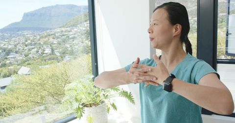 Asian woman stretching by window wearing smartwatch, gazing at mountain valley view