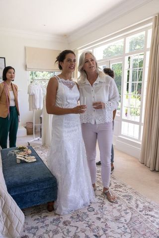Bride trying on lace wedding gown with mother celebrating with friends in sunlit garden room