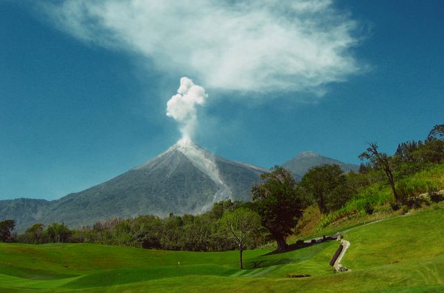 This image displays an active volcano with smoke billowing against a clear blue sky, set in a serene countryside landscape. The green fields and winding path in the foreground contrast with the dramatic volcanic activity. This image is ideal for use in travel brochures, geological studies, nature magazines, or educational materials about volcanology.