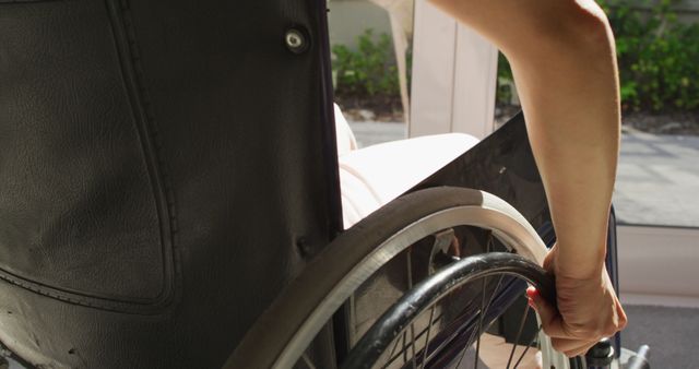 Person is actively using a wheelchair by turning wheel with hand, in outdoor area. Useful for illustrating themes of independence, support for disabilities, and accessibility in public spaces.