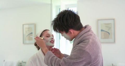 Diverse couple applying facial masks during skincare routine in bright minimalist bathroom