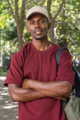 Confident african man standing arms crossed wearing maroon shirt and cap in sunlit park